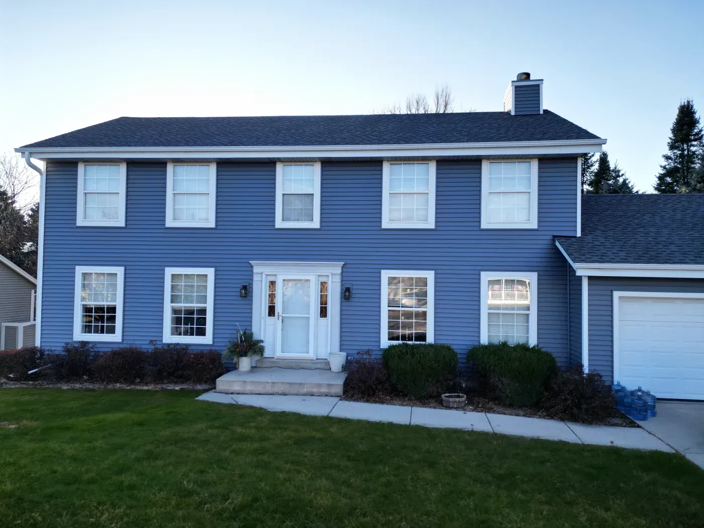 Two-story blue vinyl sided house with white trim windows, white front door, attached garage, and dark roof
