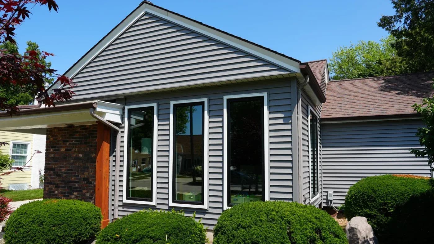 Modern house with gray horizontal siding, gabled roof, large front windows, orange entry door, and manicured green shrubs in the front yard
