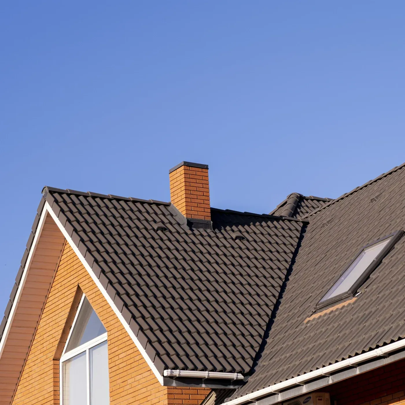 Close-up of a residential roof with dark tile roofing, brick chimney, and skylight beneath a clear blue sky.