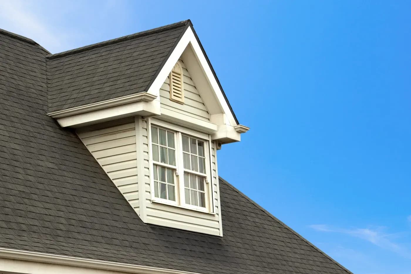 White dormer window with multi-pane glass protruding from dark slate roof with ventilation louver in gable peak against clear blue sky