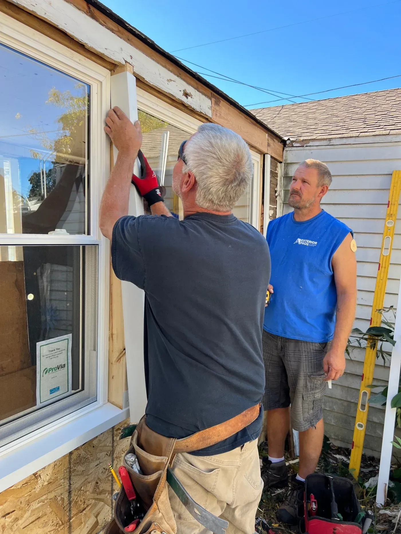 Contractor installing window frame on house exterior while coworker observes, with tools and ladder visible