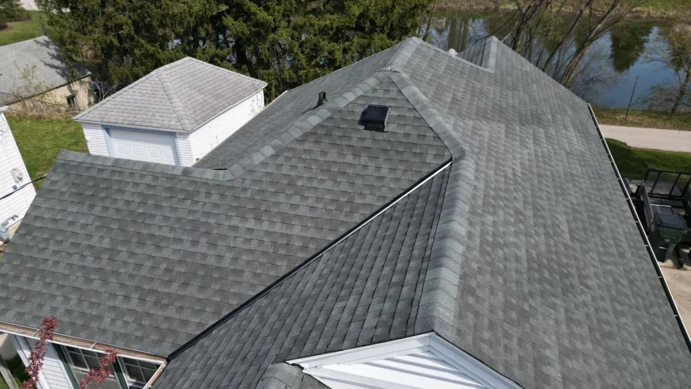 Aerial view of a house with gray architectural shingle roof and metal valley flashing, adjacent garage with peaked roof, and waterfront wooded landscape in background