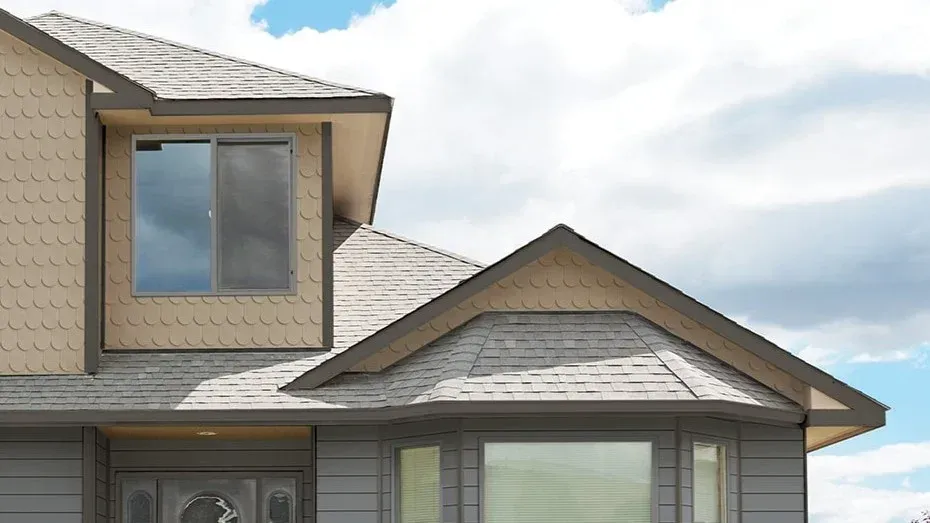 Two-story residential home featuring a peaked roof with gray shingles, tan siding with scalloped details, upper-story bay window with dark frame, and lower-level entrance with glass door