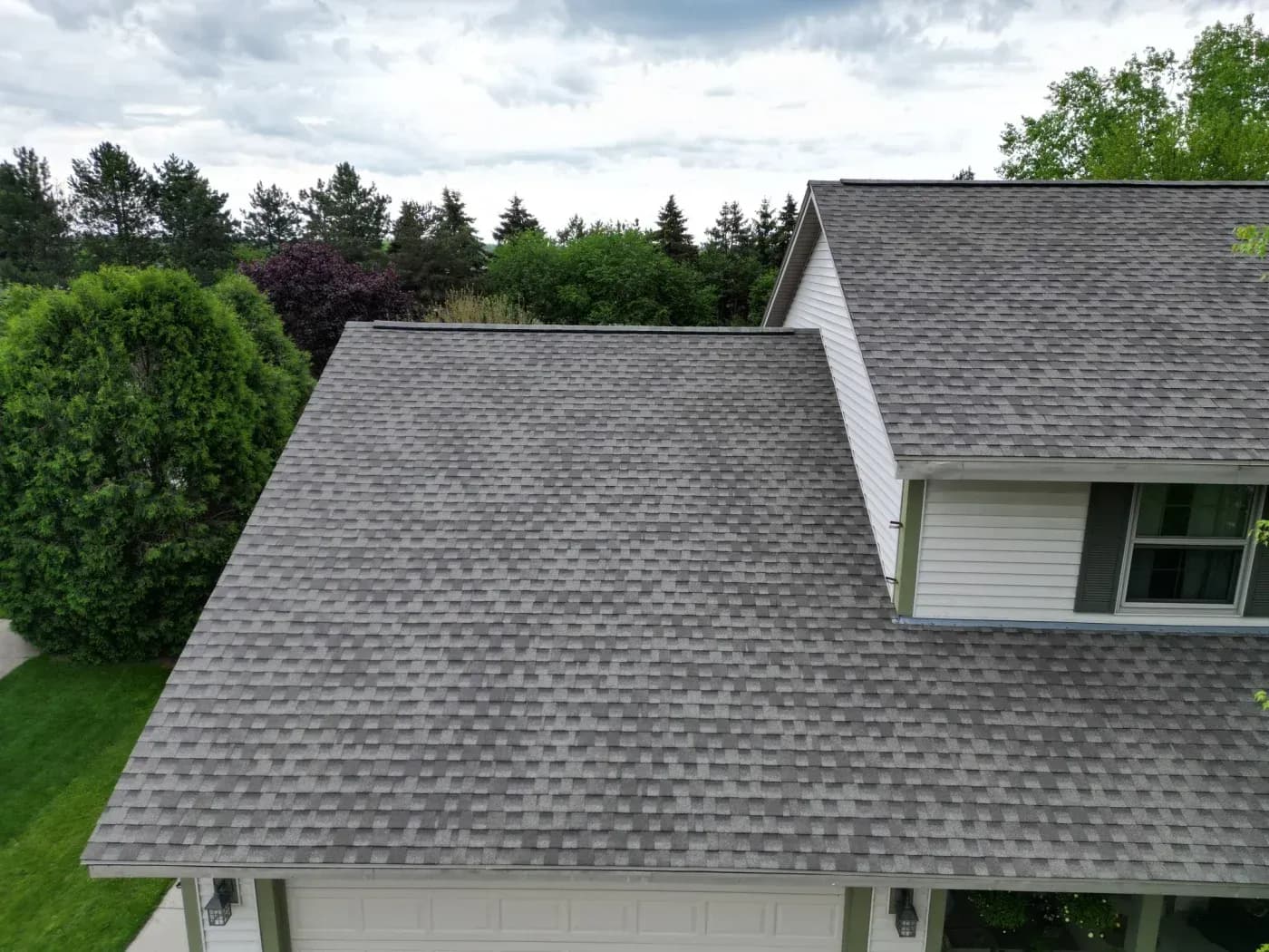 Residential house with gray shingle roof and white siding, surrounded by green landscaping and evergreen trees under cloudy sky