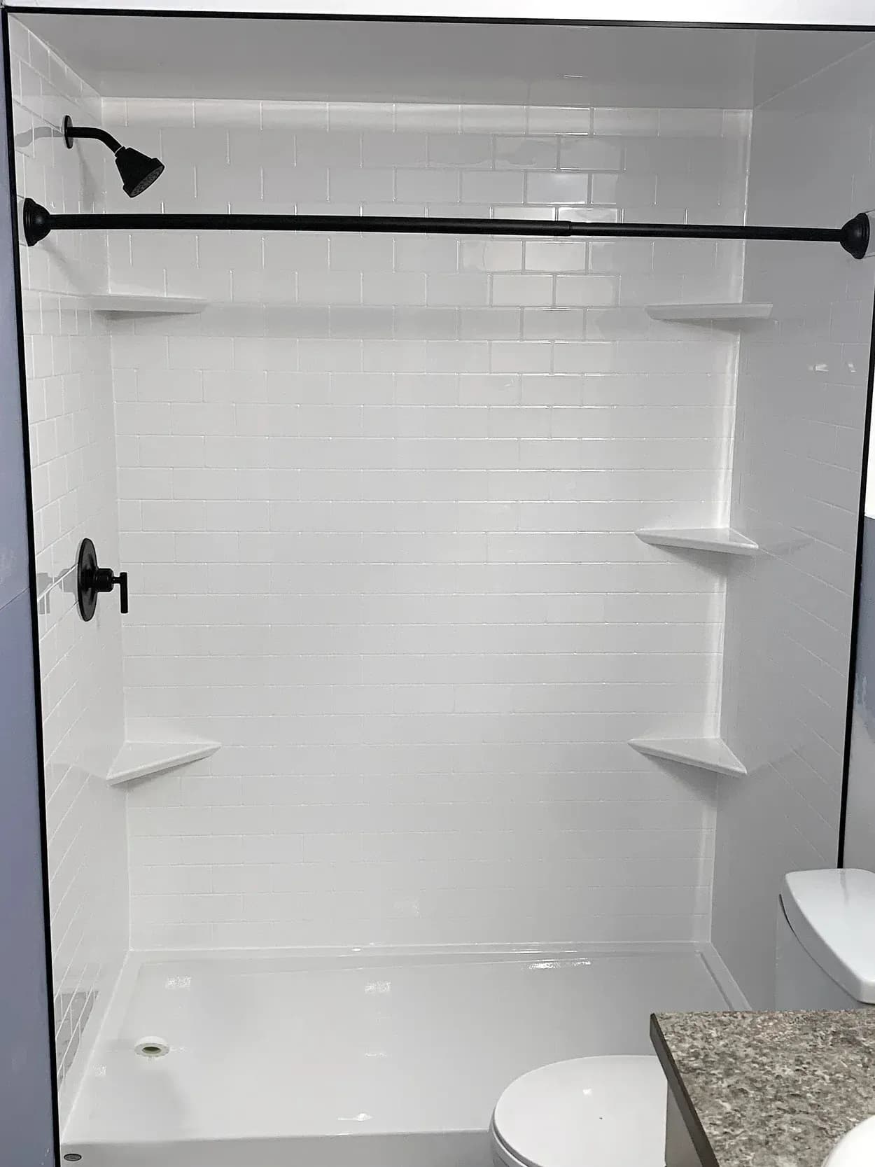 Modern bathroom shower enclosure with white subway tile walls, built-in shelving, black showerhead and fixtures, and glass door, adjacent to toilet