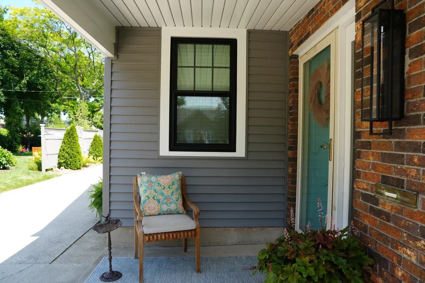 Covered porch with wooden chair, black-framed window, dark shutters, green door, and red brick wall adjoining a modern gray siding exterior