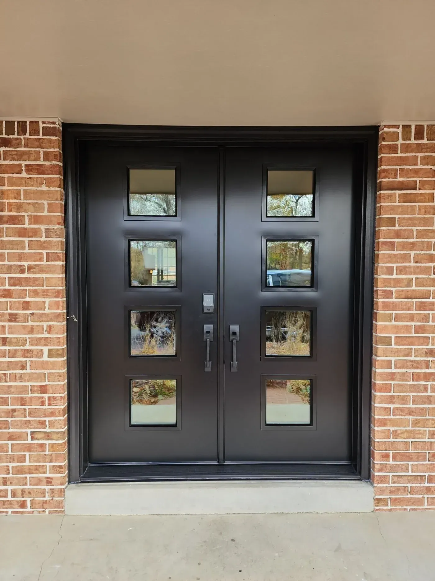 Black double entry doors with eight square windows set in red brick exterior, flanked by brick pillars