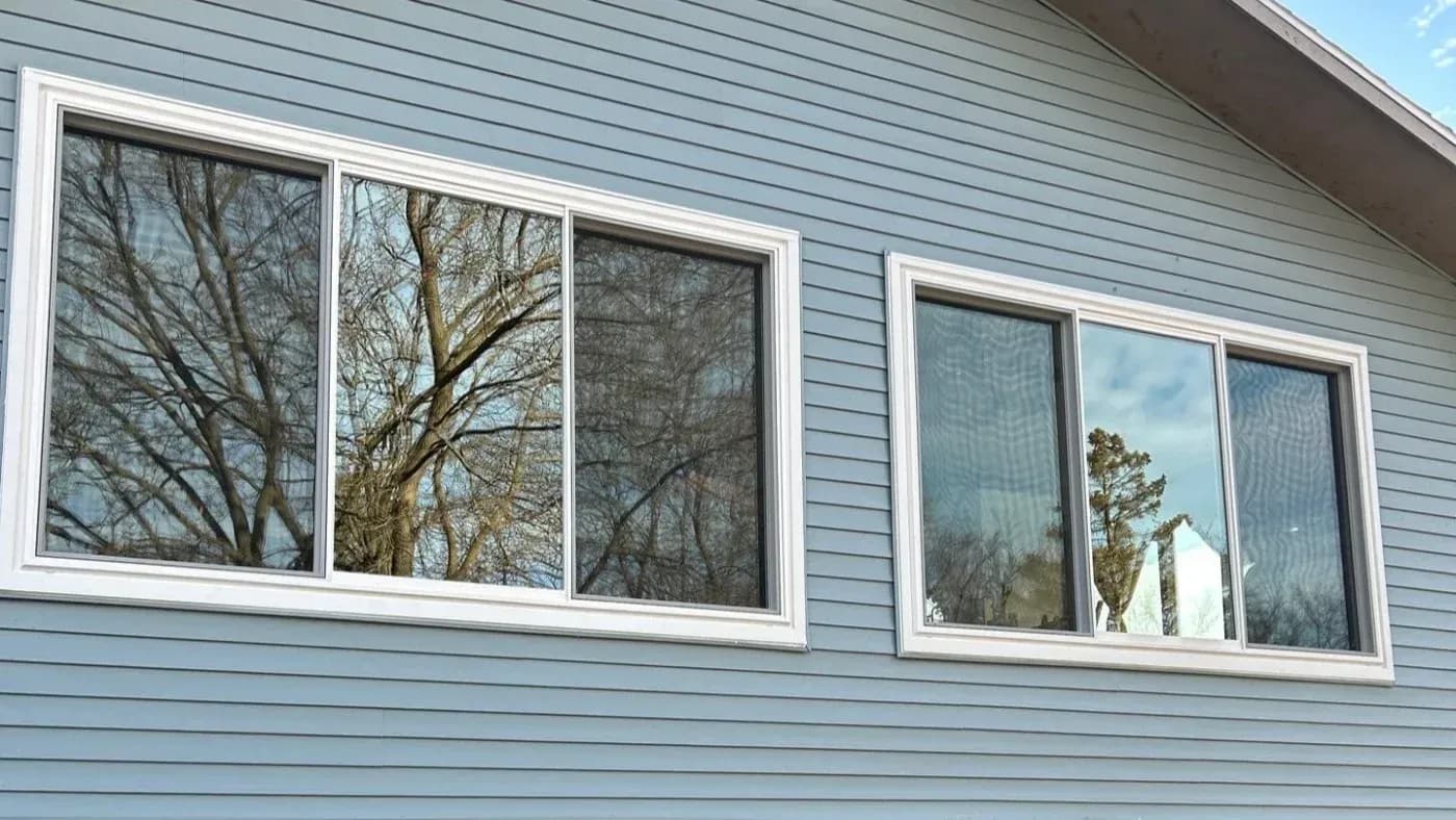 Row of five white-framed windows with white trim on light blue vinyl siding, reflecting bare trees and sky
