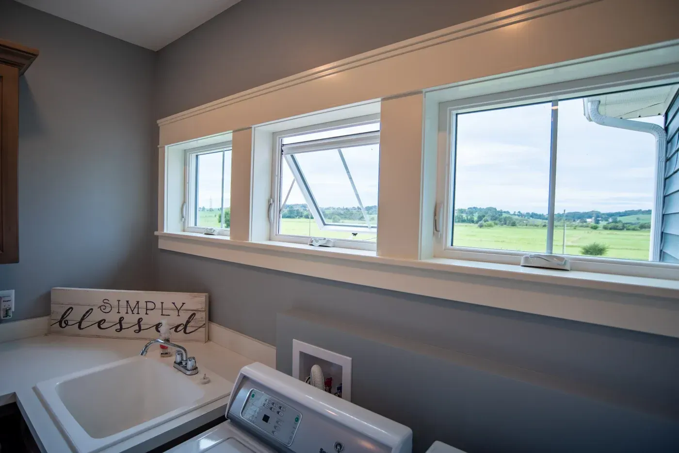 Bathroom with white sink and decorative "Simply blessed" sign, large windows overlooking rural green fields and countryside landscape