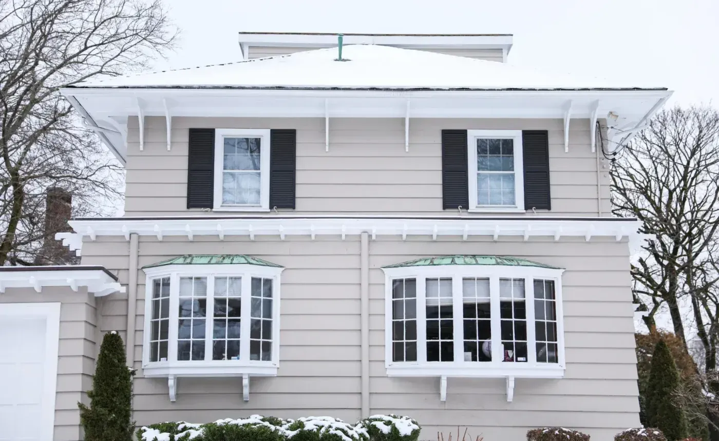 Two-story home exterior with white bay windows, black shutters, and snow on the roof.
