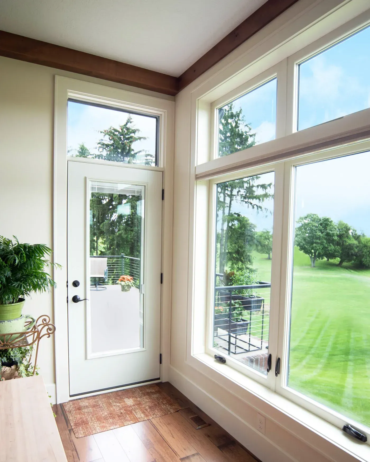 Bright sunroom with white door, expansive windows overlooking manicured lawn and trees, potted plants on left, wooden beams on ceiling