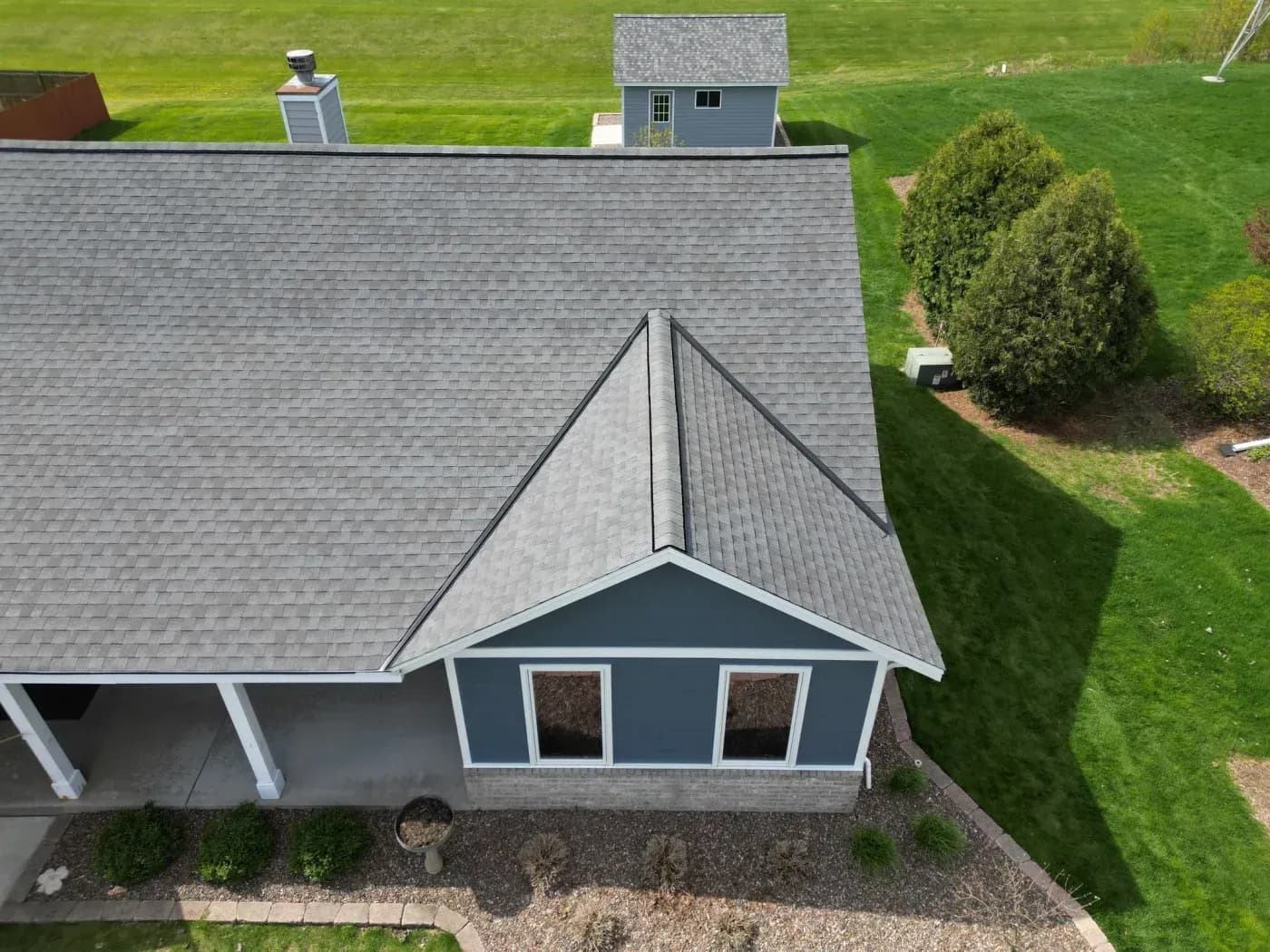 Aerial view of residential home with gray shingle roof, dark blue addition with peaked roof and two windows, attached garage, and manicured lawn with evergreen shrub