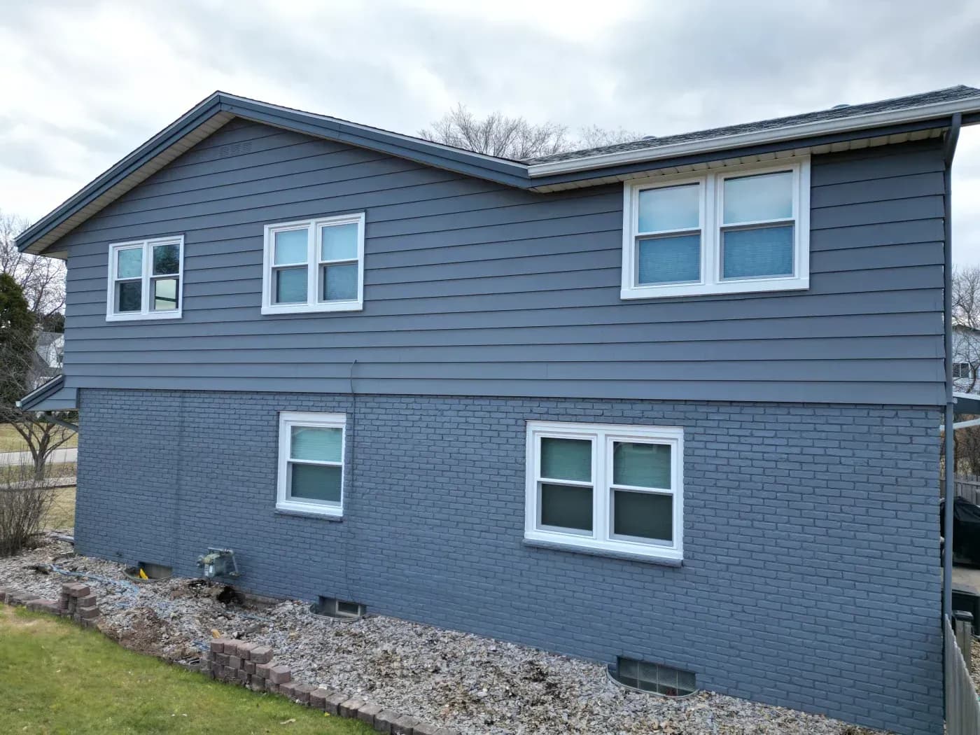 Two-story residential house with blue siding on upper level and blue brick on lower level, featuring white-trimmed windows and gravel foundation landscaping