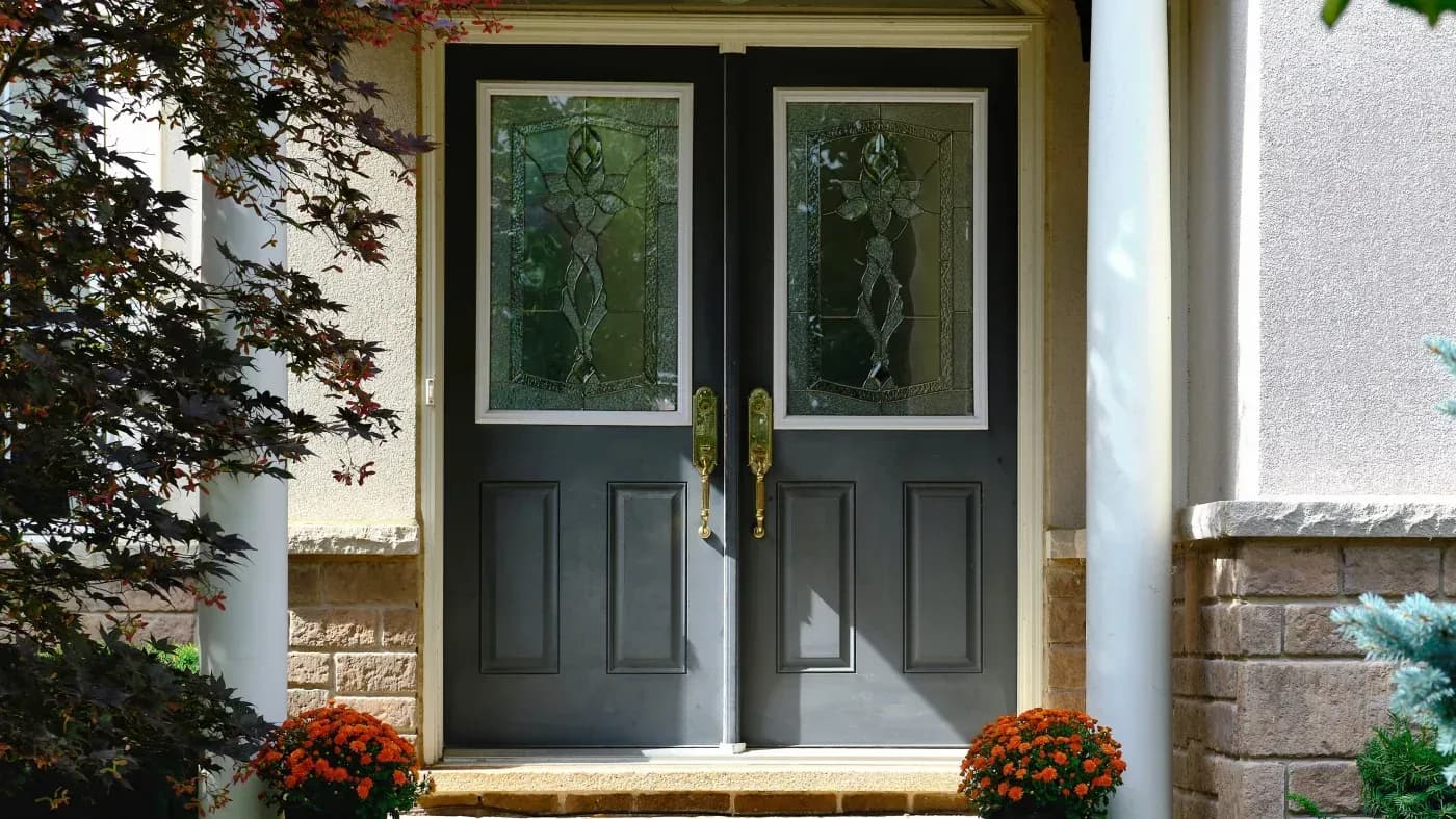 Pair of black double doors with decorative glass panels featuring leaf designs, flanked by orange mums and stone columns on a house entrance
