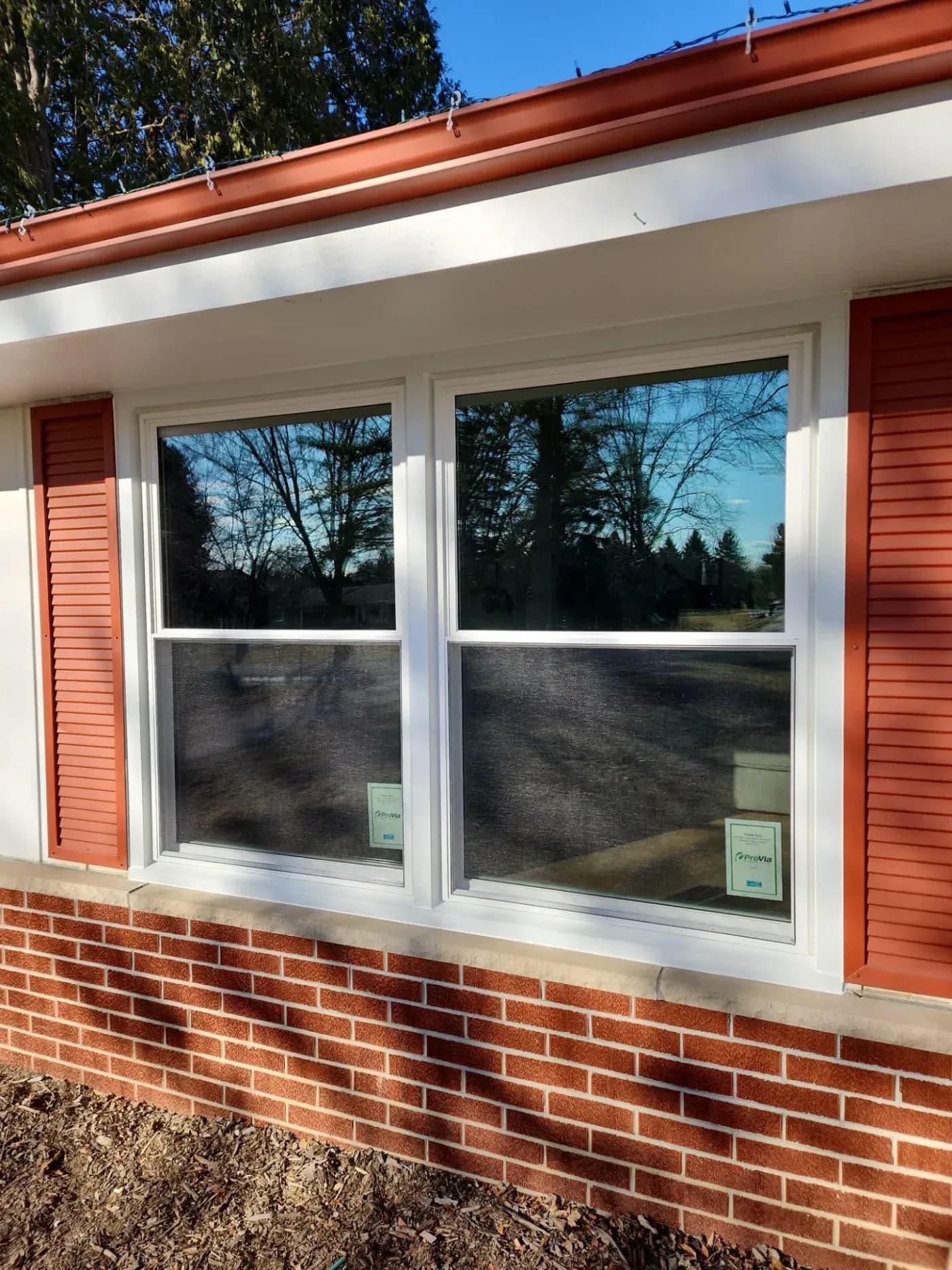 Two white double-hung windows with screens on a brick house exterior, with red shutters and copper gutters, overlooking bare trees and clear sky
