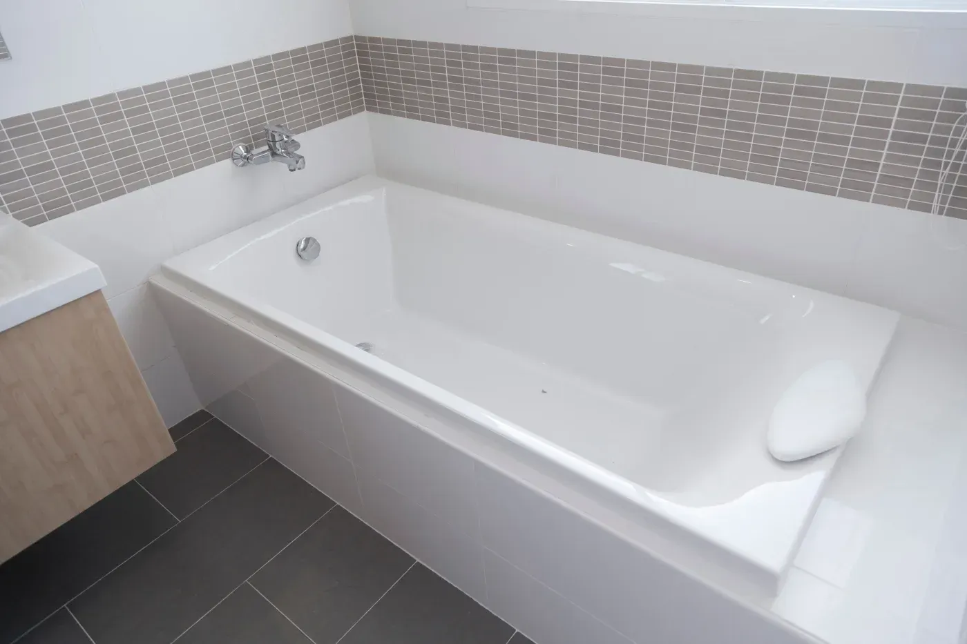 White bathtub with a built-in headrest, gray mosaic tile border, and dark floor tiles.