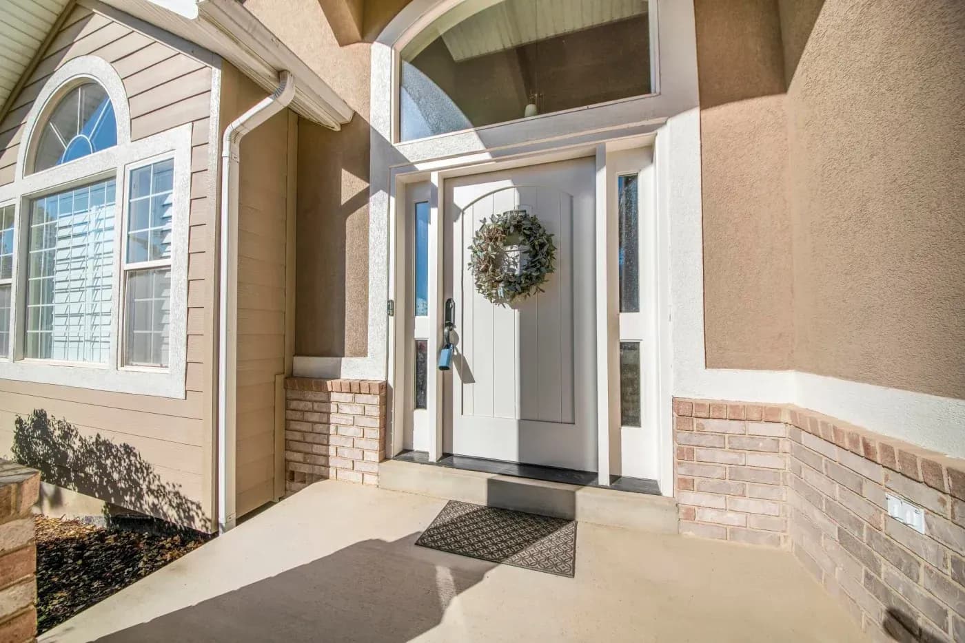 Home entrance with white front door decorated with a green wreath, flanked by brick walls and windows, with a welcome mat on the concrete porch
