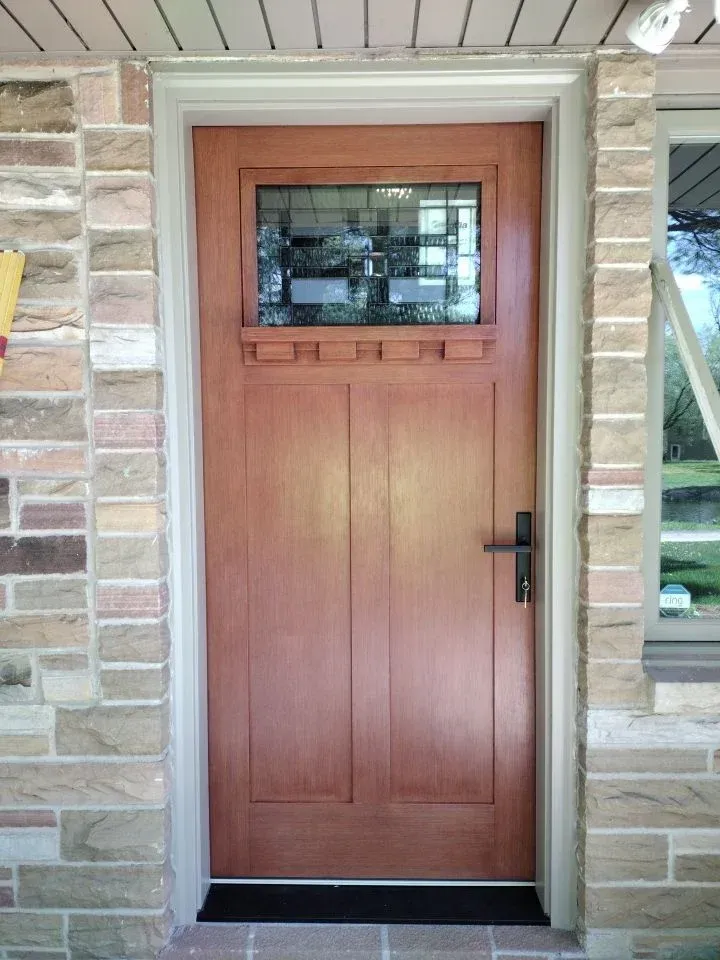 Red-brown wooden front door with small window and black handle, set in stone frame on residential entryway