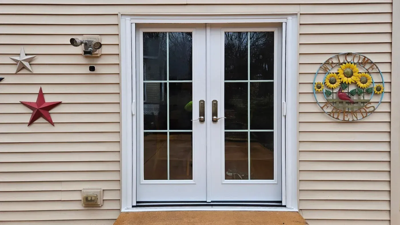 White double-pane patio doors with grid pattern on tan siding, flanked by red star decoration on left and sunflower wreath on right, with motion light above and doorbell below