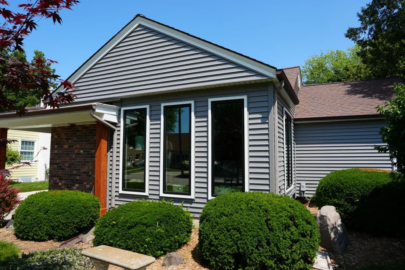 Contemporary house addition with gray horizontal siding, large glass doors, triangular roof peak, surrounded by green shrubs and landscaping