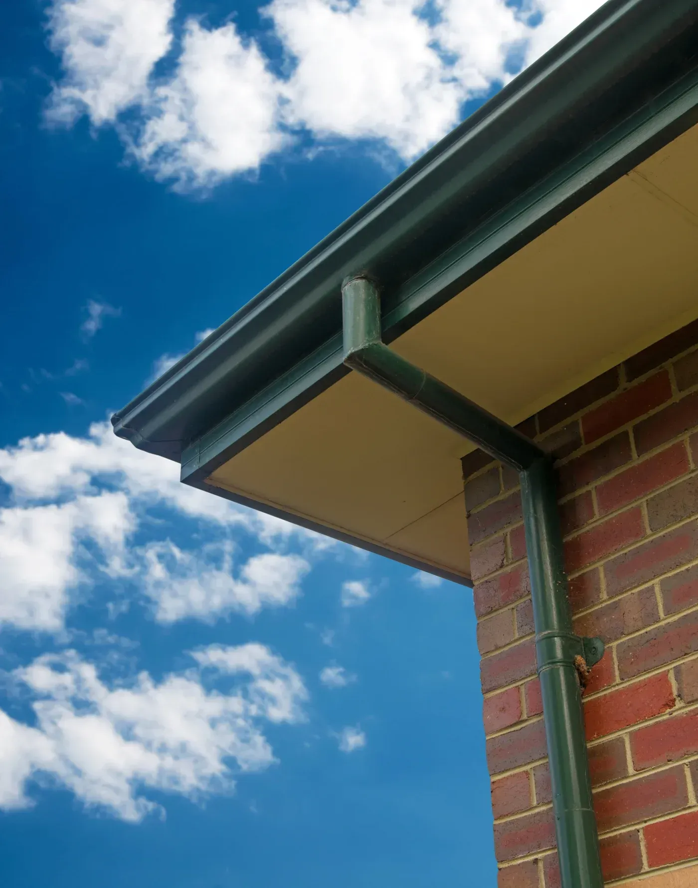 Green metal fascia and downpipe on brick building corner against blue sky with white clouds