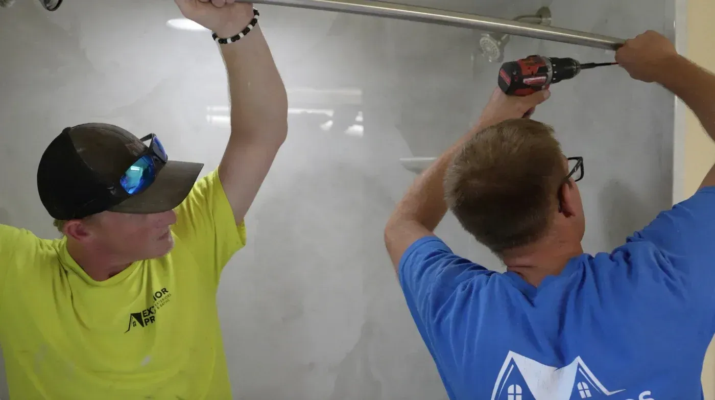 Two workers in safety gear installing drywall on a ceiling using power tools