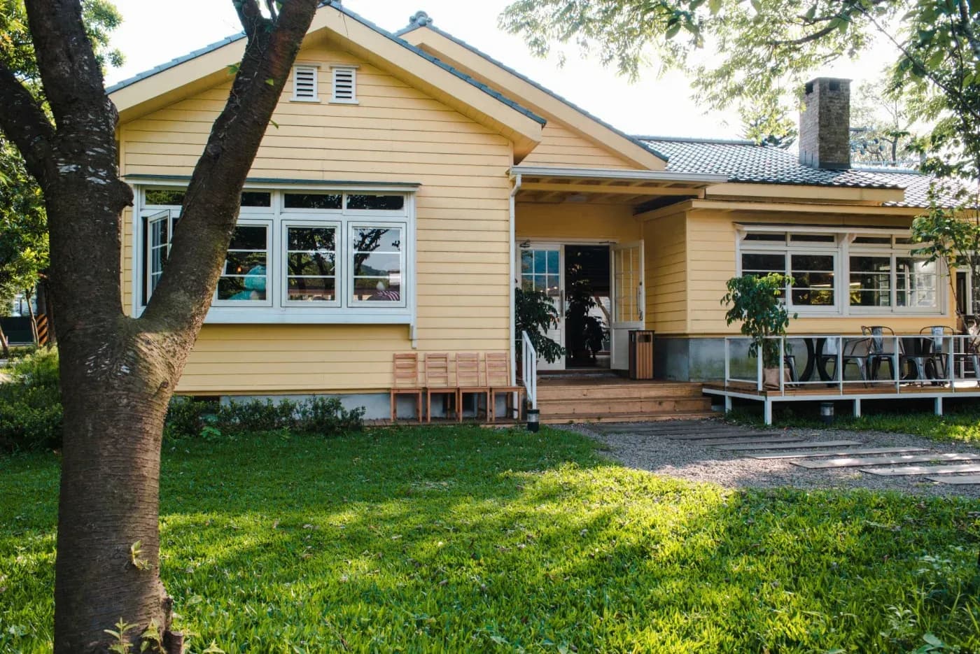 Cream-colored residential home with blue roof, large windows, wooden decks, and mature tree in front yard overlooking water