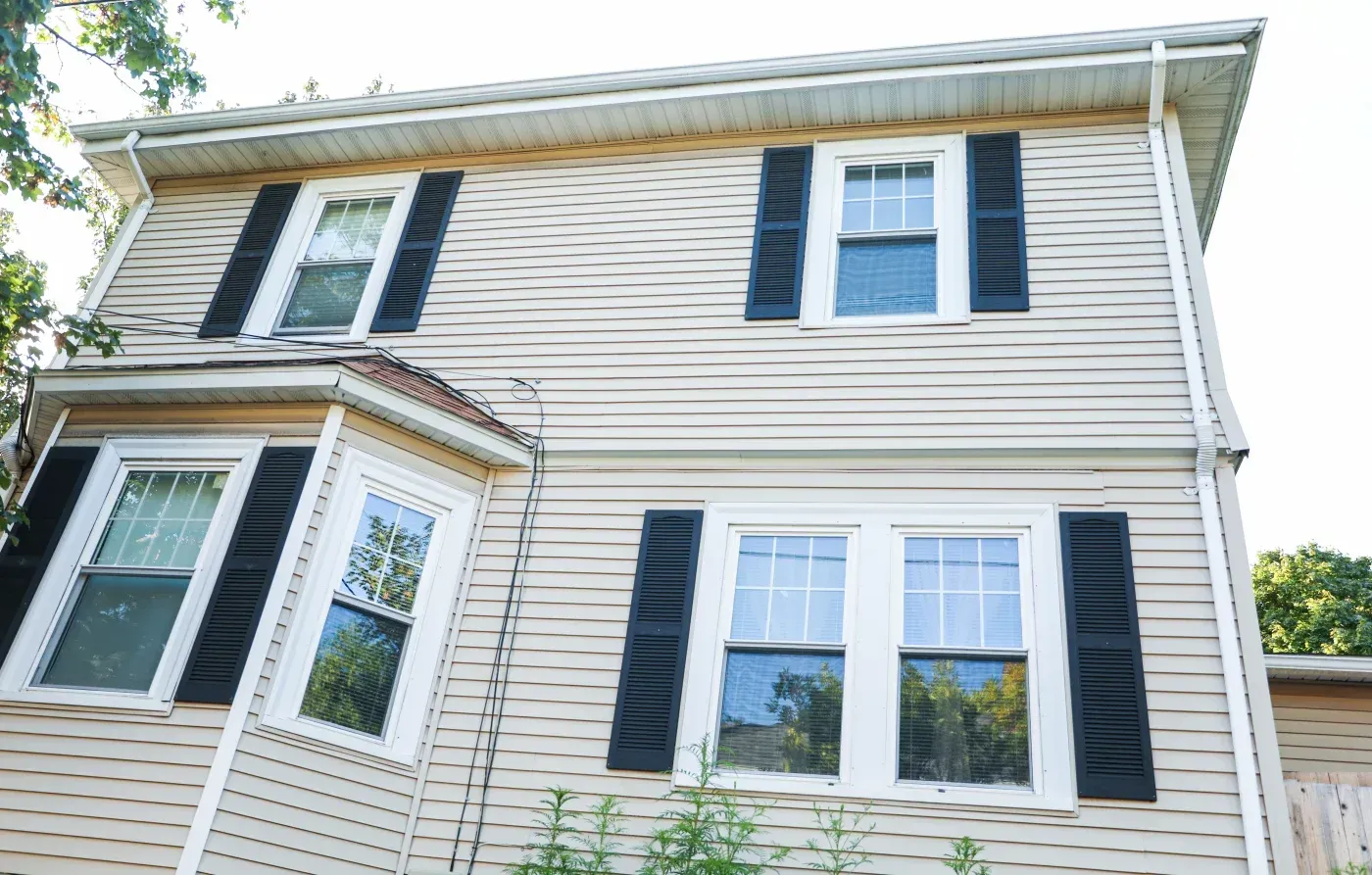 Two-story house with tan vinyl siding, black shutters, and white-trimmed windows, featuring a side entrance and metal roof