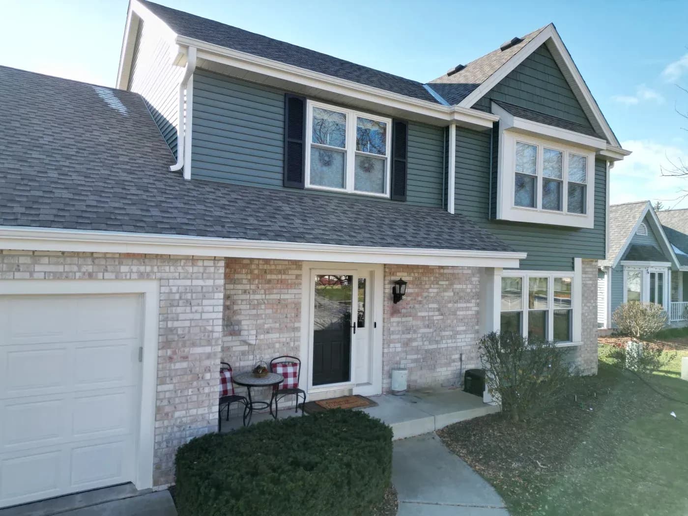 Two-story suburban house with green and white siding, brick lower level, front patio with seating, garage on left, and dormer windows on upper floor