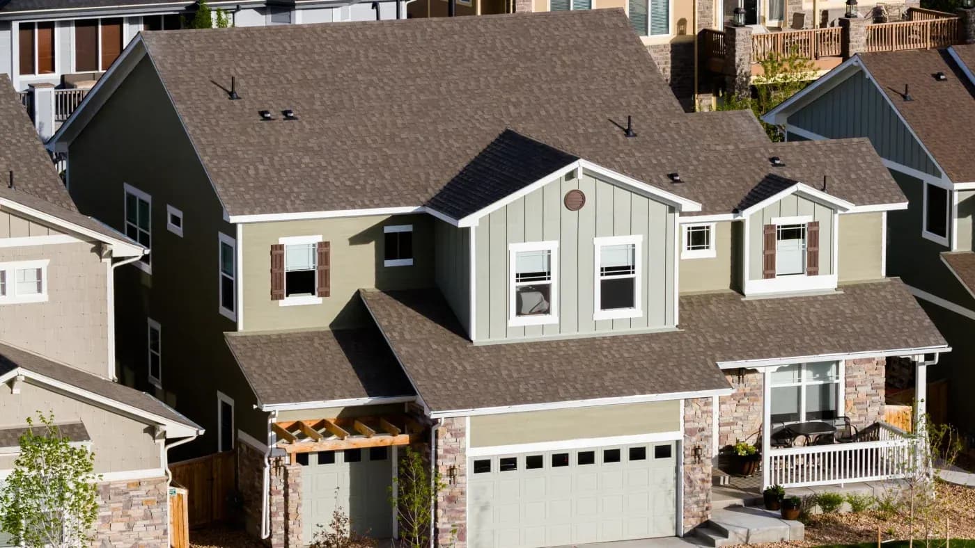 Suburban neighborhood street view showing multiple residential homes with brown shingled roofs, varying exterior colors including cream, gray, and dark siding, white-trimmed windows, and brick or stone accents