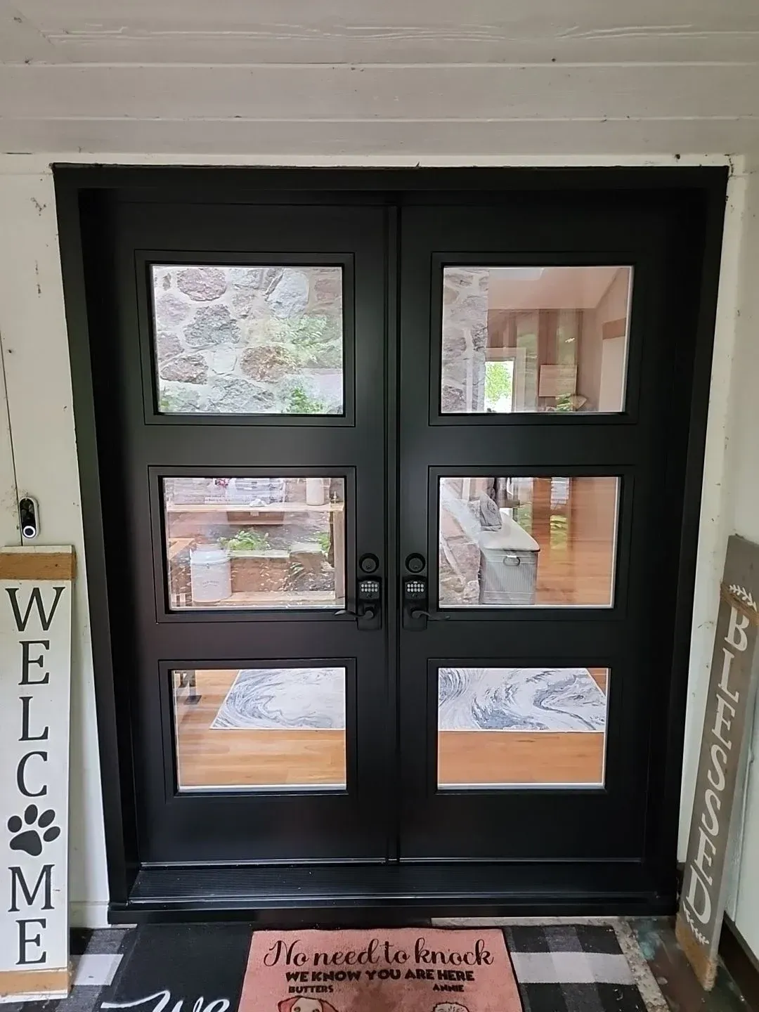 Double black entry doors with six glass panes showing interior and exterior views, flanked by welcome signs and a doormat reading "No need to knock we know you are BUTTERS