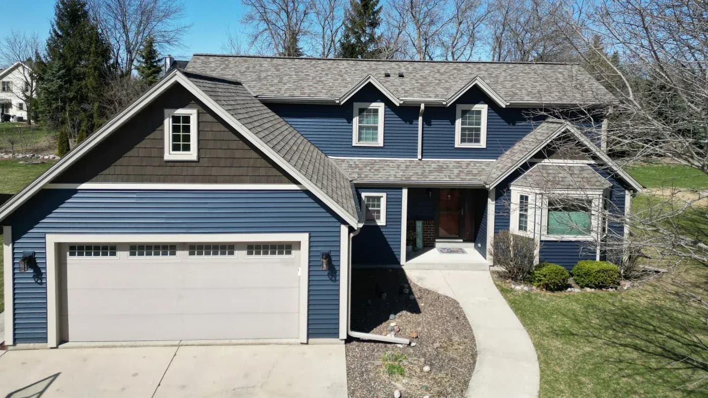 Blue and gray two-story residential house with attached two-car garage, three dormers, concrete driveway, and landscaped front yard with evergreen trees