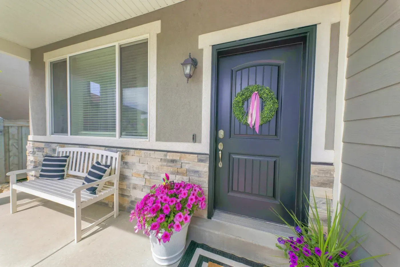 Front porch with white bench, dark blue door adorned with green wreath and pink ribbon, bright pink petunias in planter, and striped outdoor cushions