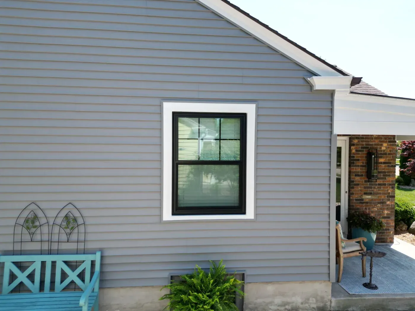 House exterior with gray horizontal siding, black-framed window with white trim, turquoise bench on left, and brick section visible on right
