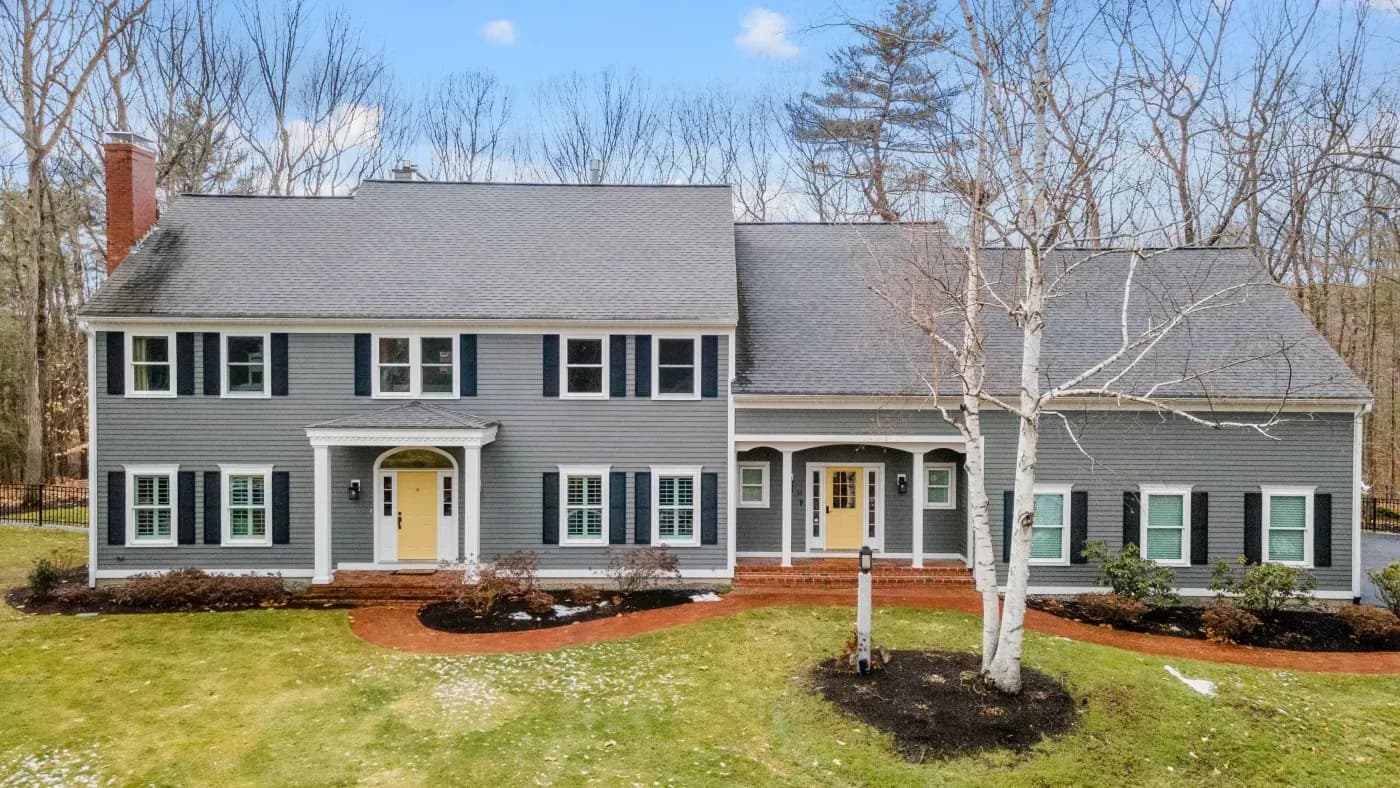 Two-story gray colonial house with yellow doors, black shutters, brick chimney, and attached garage, surrounded by bare trees and manicured lawn