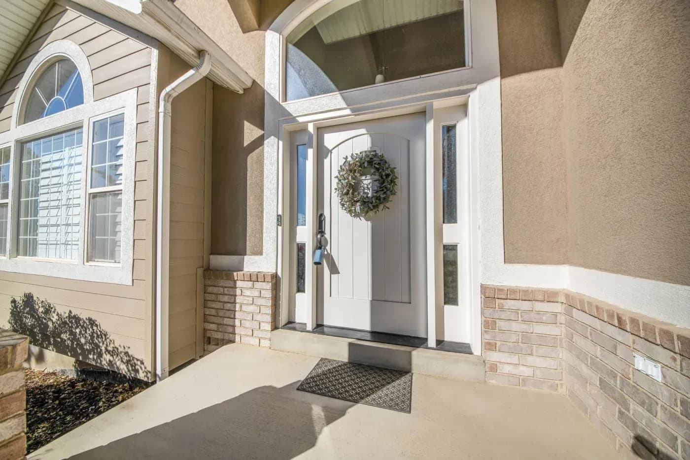 Front entry of a home featuring a white door with a green wreath, flanked by windows and brick pillars, with a concrete porch and welcome mat