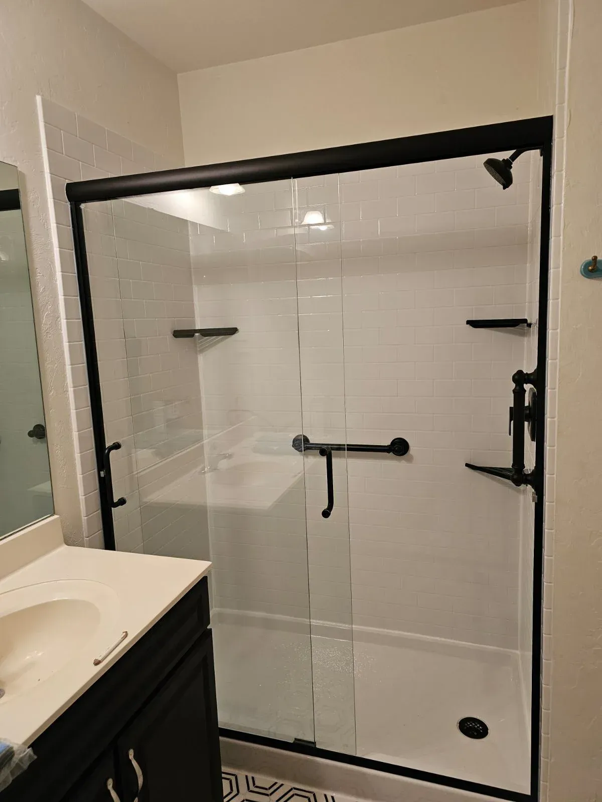 Shower enclosure with black frame and glass doors, white subway tile walls, black grab bars, and rainfall showerhead mounted in modern bathroom