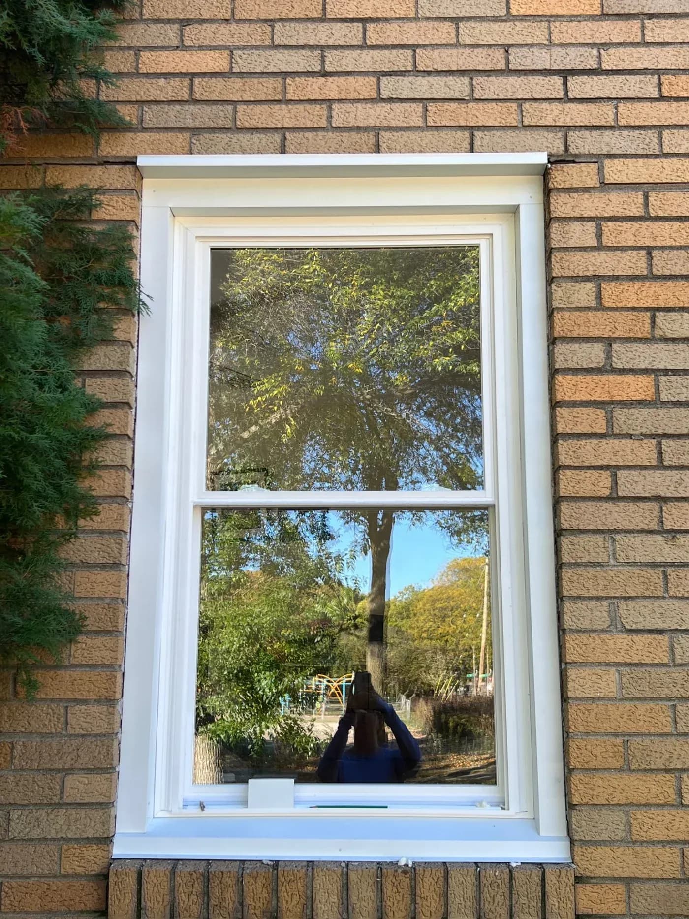 White-framed window in brick wall reflecting trees, sky, and a person's silhouette against outdoor garden landscape