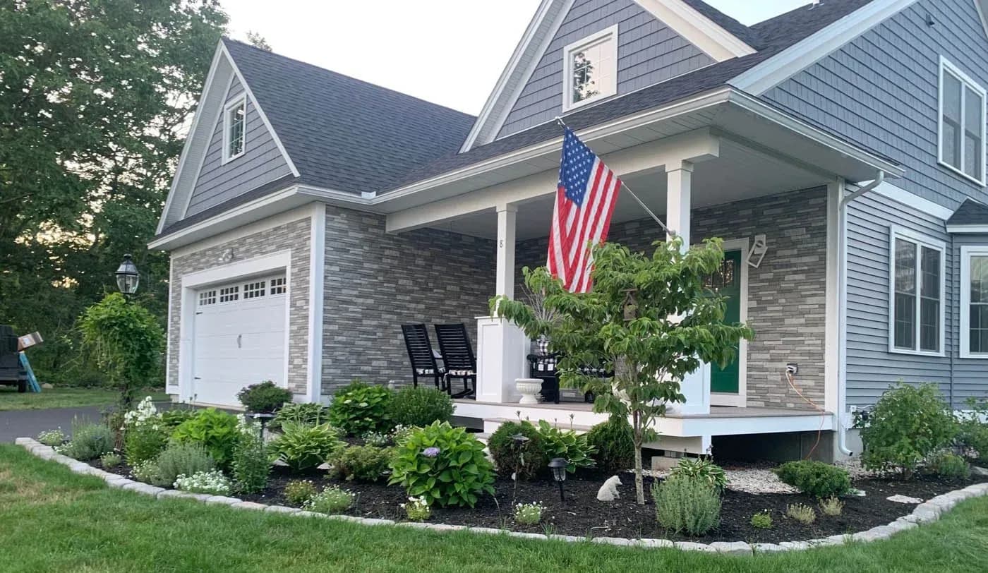 Contemporary two-story home with brick and siding exterior, white columns on front porch, American flag hanging from porch, landscaped front yard with shrubs and plants, attached garage on left side