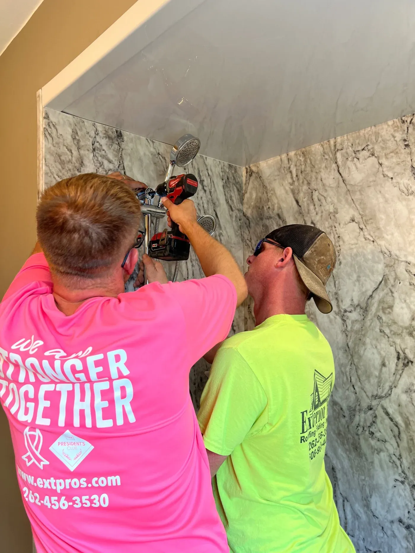 Two plumbers installing a shower head and valve on a marble tile wall during bathroom renovation
