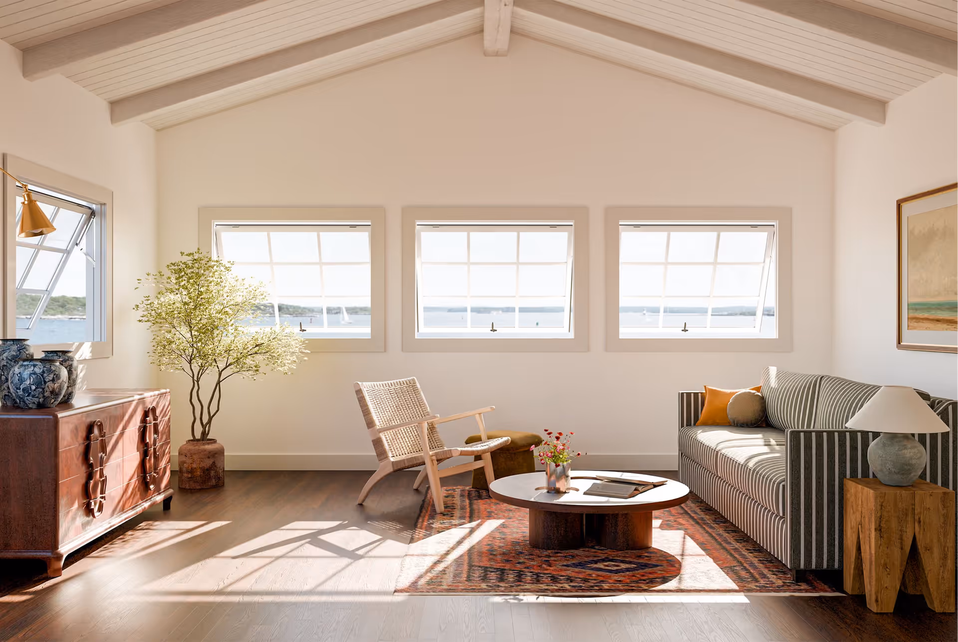Interior view of a bright, vaulted-ceiling sunroom featuring a row of three Marvin Ultimate push-out awning windows with grid panes, open to reveal a coastal water view. The space is styled with dark hardwood floors, a woven lounge chair, a striped sofa, a low wooden coffee table over a pattern rug, and a potted olive tree.