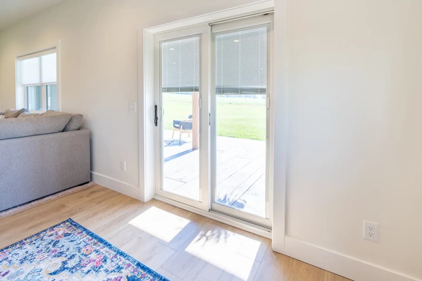 Bright living room with white sliding glass doors opening to a patio and grassy yard, featuring a gray sofa, blue patterned rug, and wooden flooring