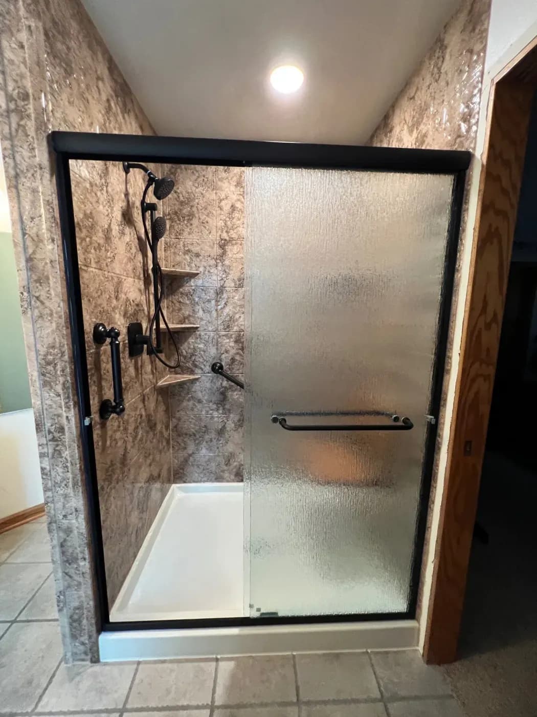 Shower enclosure with black frame, textured glass door, and stone tile walls with handheld showerhead and shelves inside