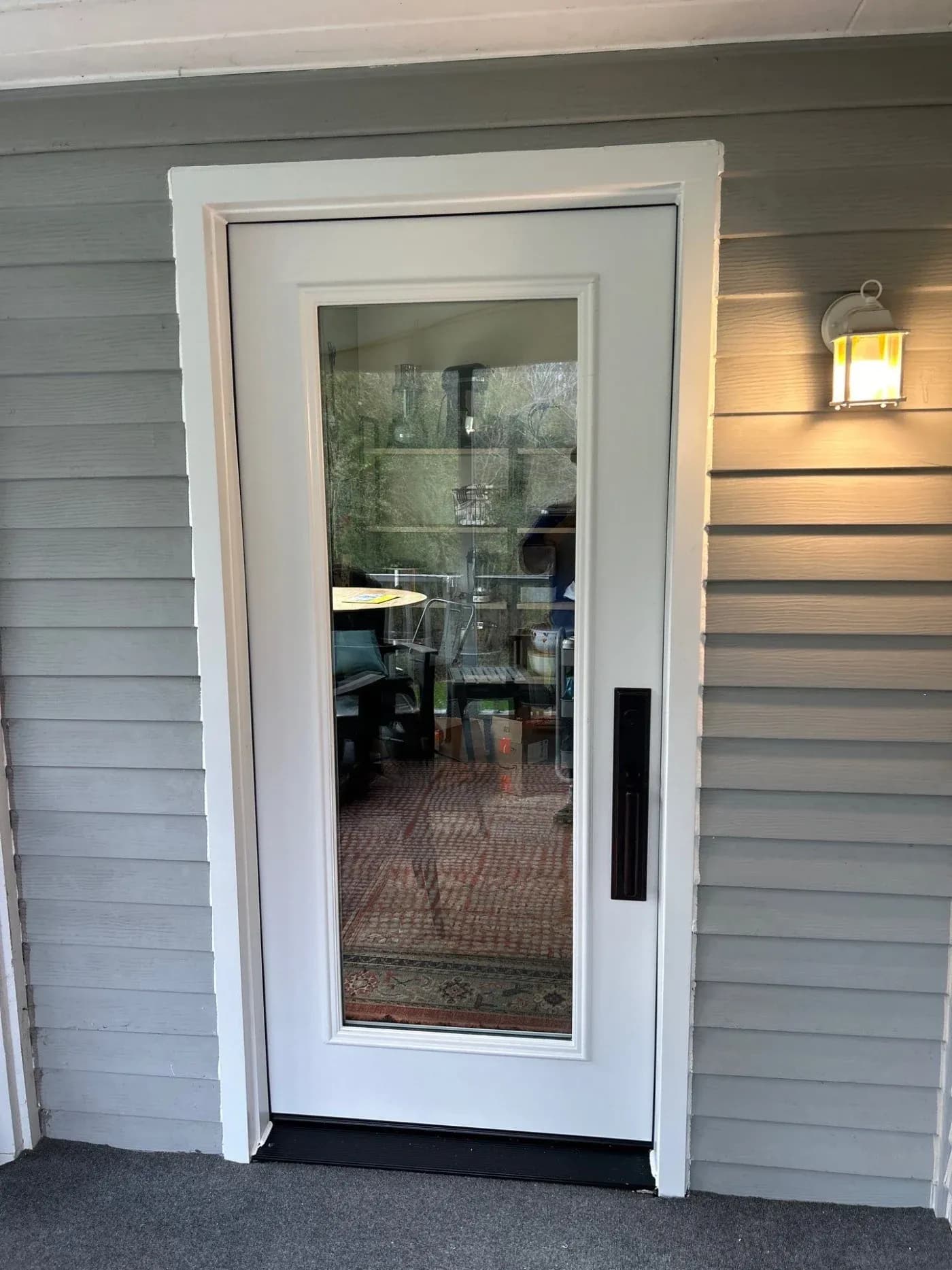 White exterior door with full-length glass panel, black handle, and transom window above, mounted on gray horizontal siding with porch light to the right