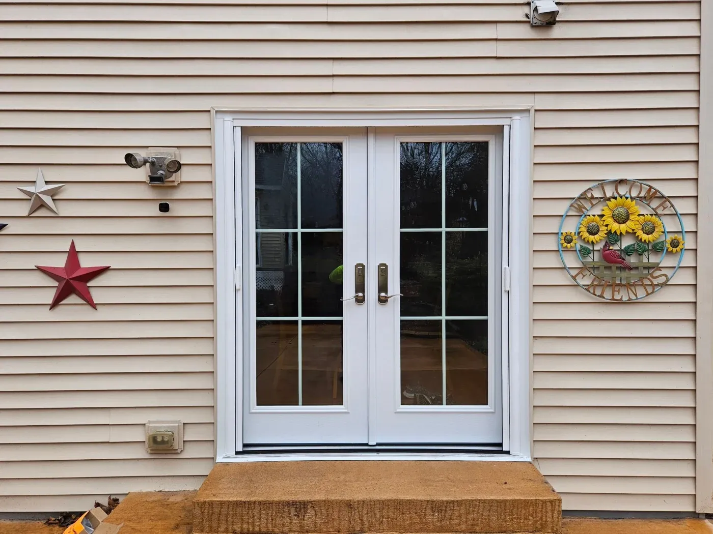 White French doors with 8 panes on tan siding, flanked by decorative red and silver stars, a sunflower wreath, and security lighting