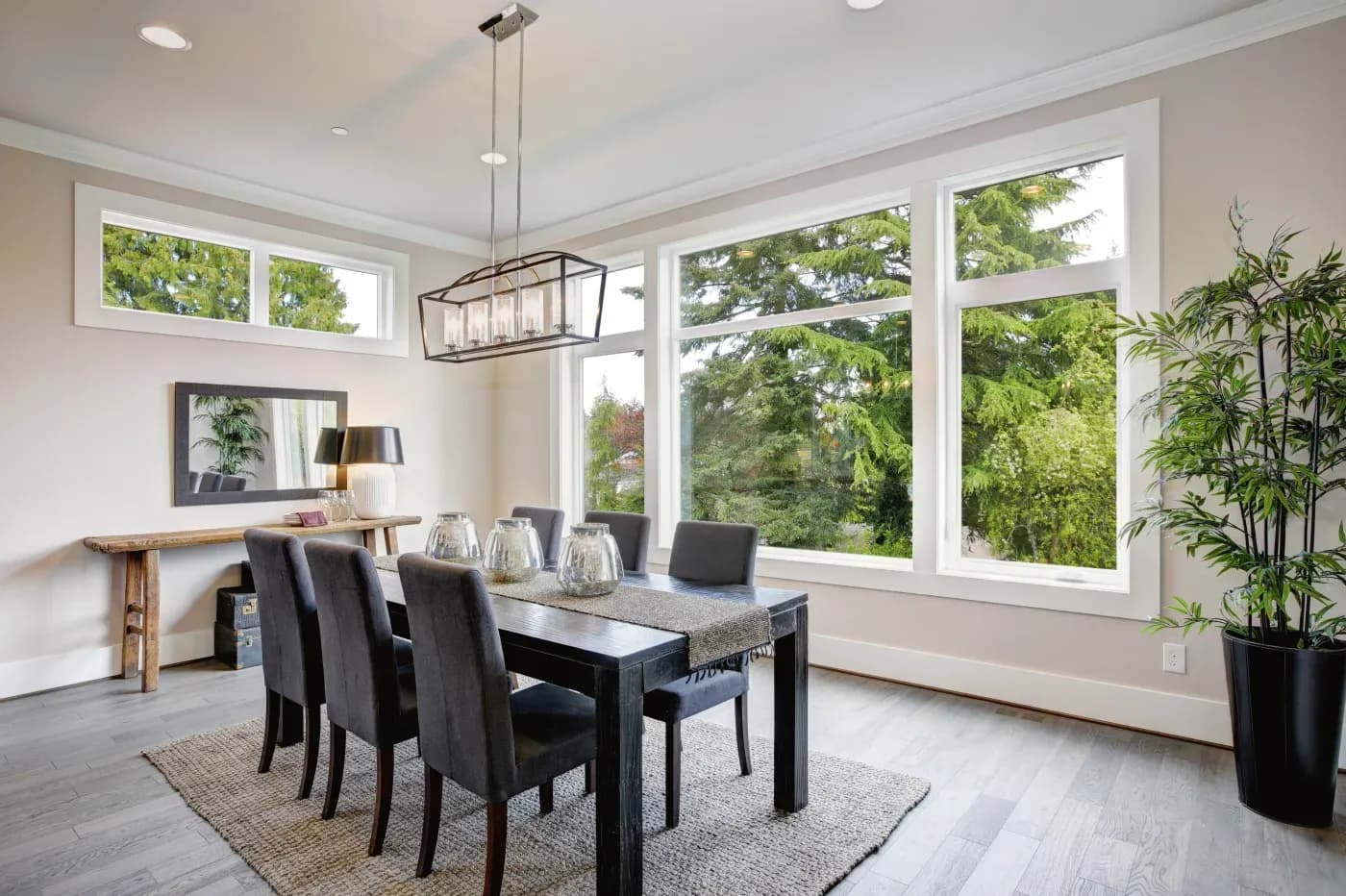 Bright dining room with black table, gray upholstered chairs, pendant light fixture, wooden console table, and floor-to-ceiling windows overlooking trees