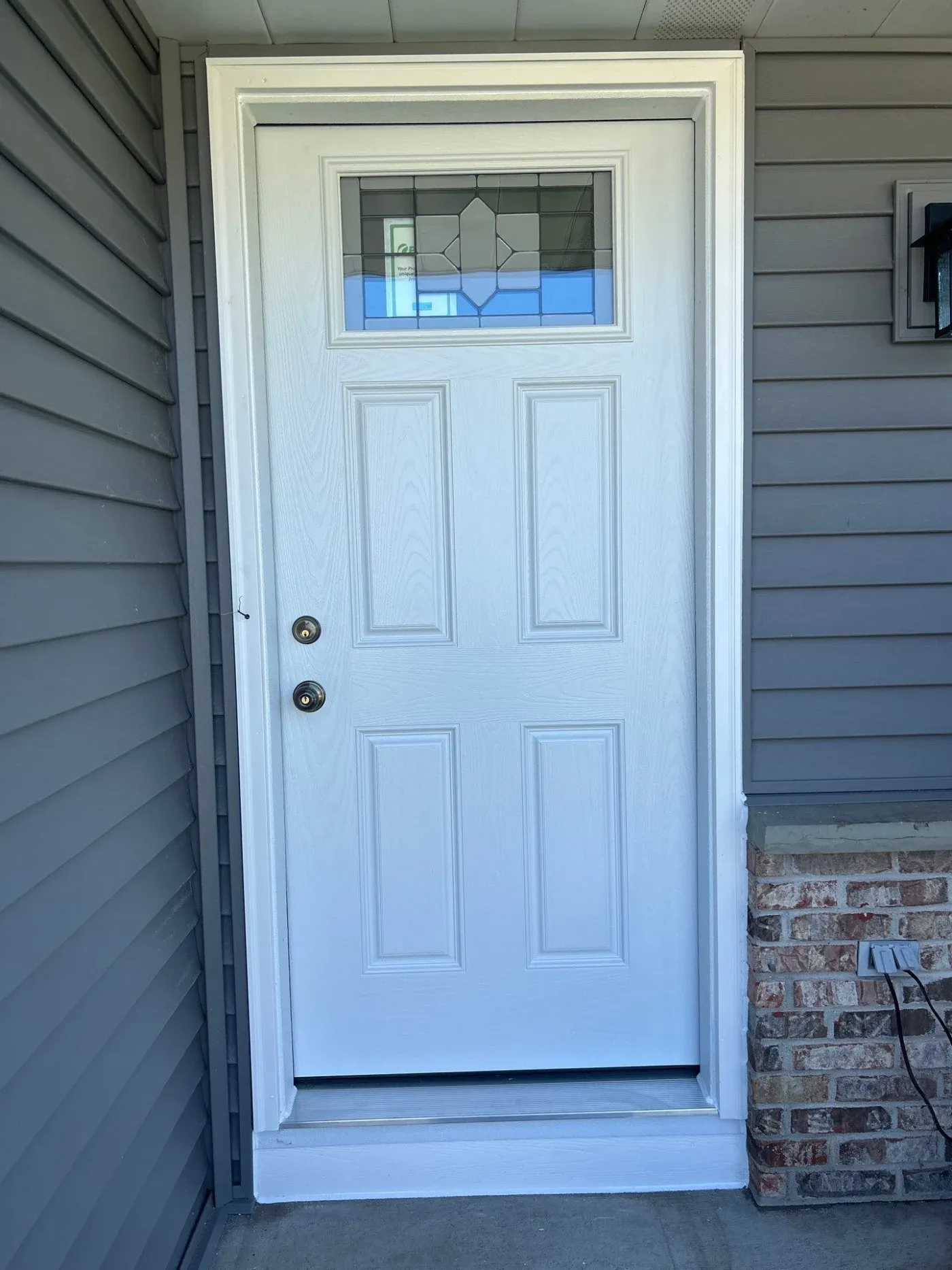 White front entry door with six panels and geometric transom window, mounted on brick and siding exterior