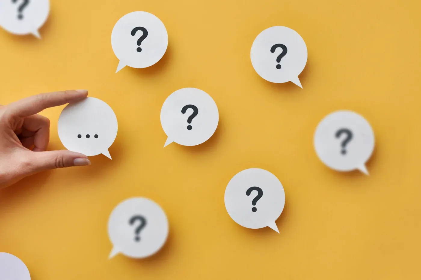 Hand holding speech bubble with three dots among multiple speech bubbles with question marks on yellow background, representing customer inquiries and support