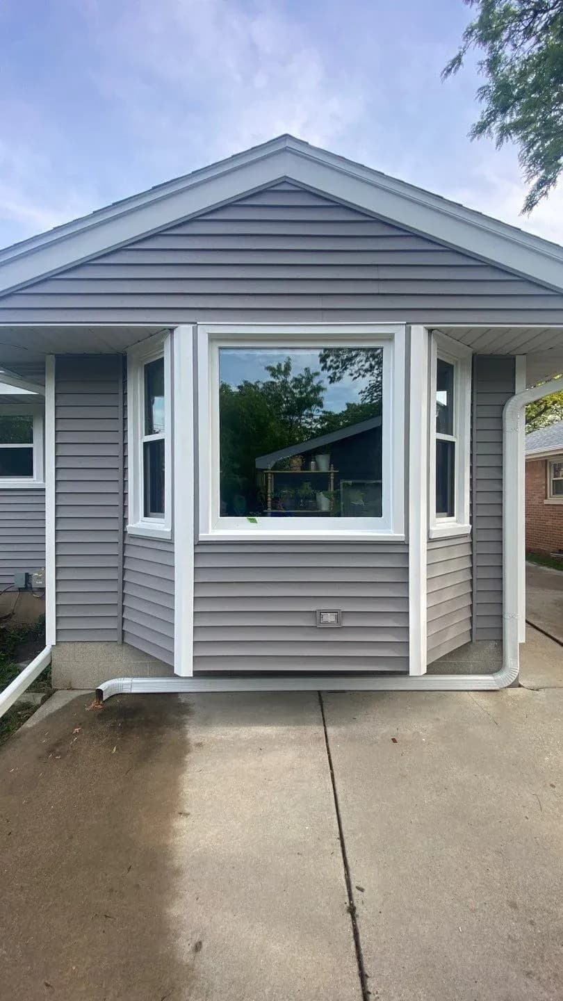 Modern gray and white residential sunroom with large central window, gabled roof, and covered side porches, viewed from concrete driveway