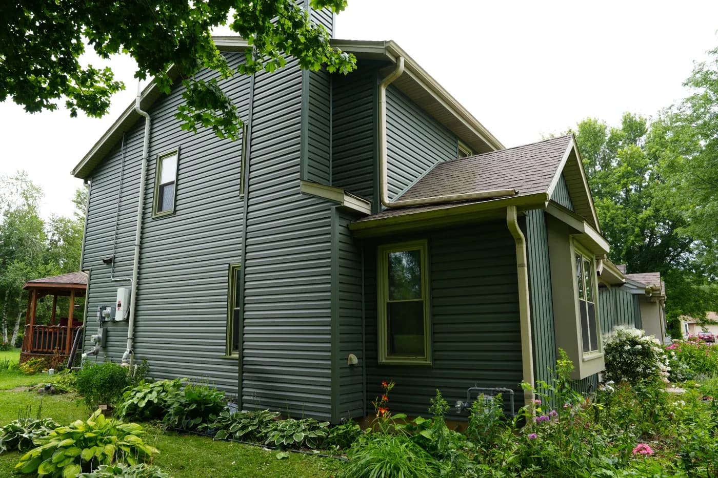 Two-story gray house with horizontal siding, green-trimmed windows and door, surrounded by mature trees and flowering garden beds with pink blooms